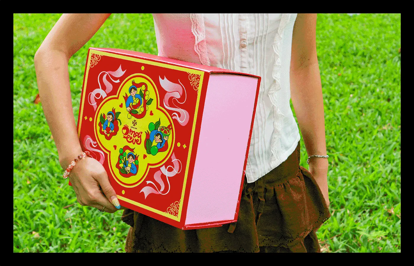 Photograph of a red Ô MAI LOVE gift box held outdoors against green grass.